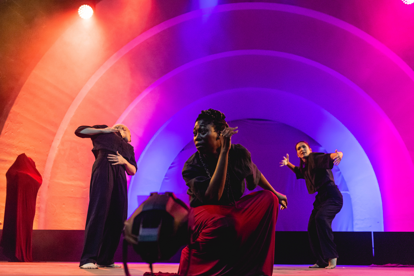 Against arches of purple, pink, and orange light, the members of Passion Fruit Dance Company groove. Downstage, one kneels while, behind her, two gesticulate.